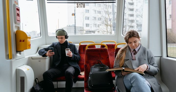 Electric Train - A Woman in Gray Coat Reading a Book while Sitting Near the Man Wearing Headphones while Holding His Mobile Phone