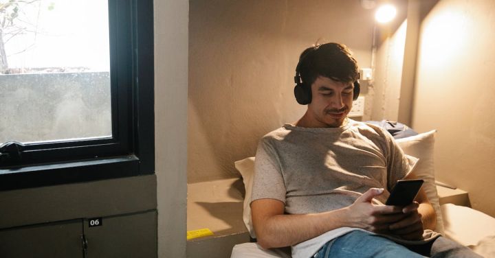 Train Music - Smiling man resting on bed and using smartphone and headphones