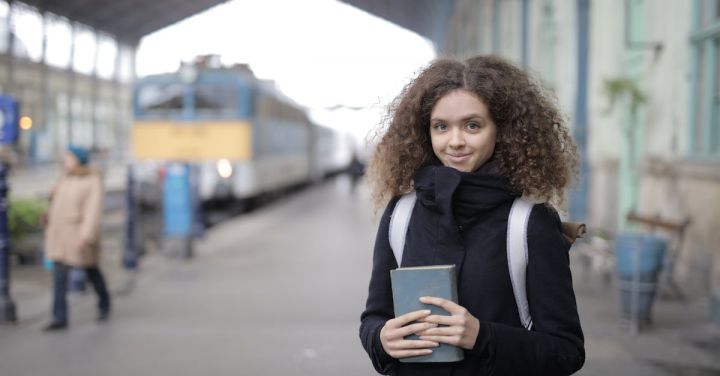 Trains In Literature - Positive young lady with backpack and book standing on platform