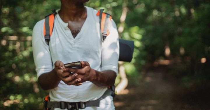 Orient Express - Crop unrecognizable African American man with backpack and smartphone searching route while going astray in green forest