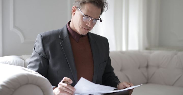 Bankruptcy Lawyer - Elegant adult man in jacket and glasses looking through documents while sitting on white sofa in luxury room