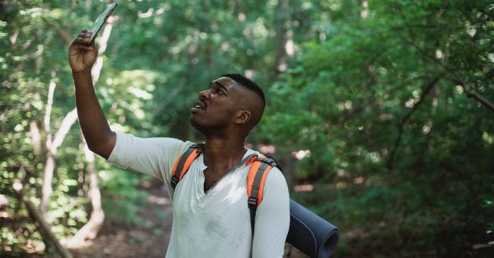 Orient Express - African American male with backpack standing in forest and holding mobile phone while catching GPS signal during hike