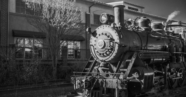 Steam Engine - Black and white fragment of old steam locomotive with chimney on railroad near building and leafless tree in city on street